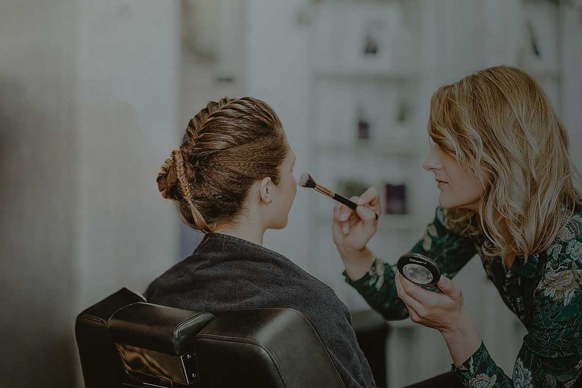 Eine Kundin, mit zu einem Zopf gestyltem Haar, bekommt ein Make-up von der vor ihr sitzenden Friseurin.